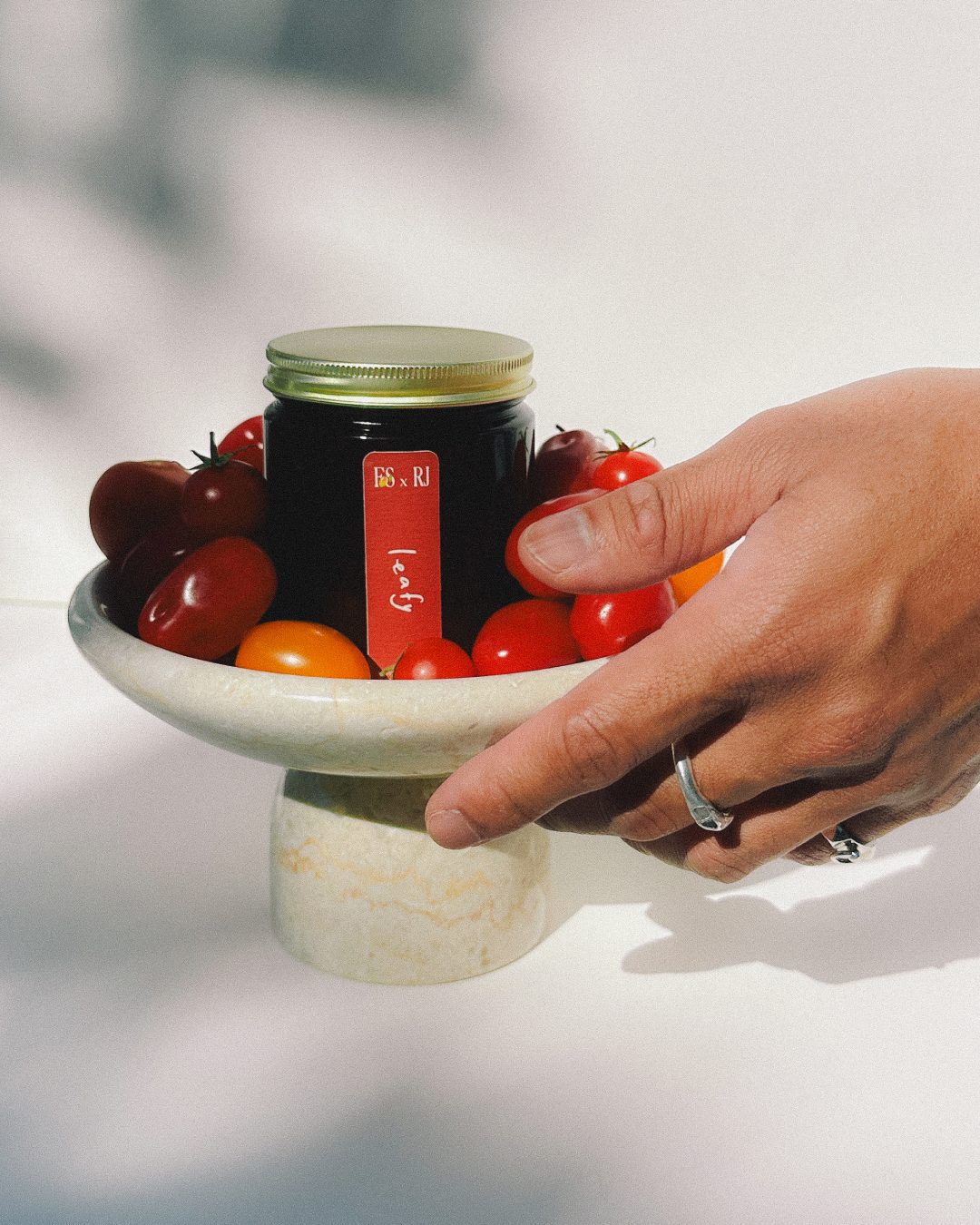 A hand reaching for a Leafy candle jar surrounded by cherry tomatoes on a marble stand